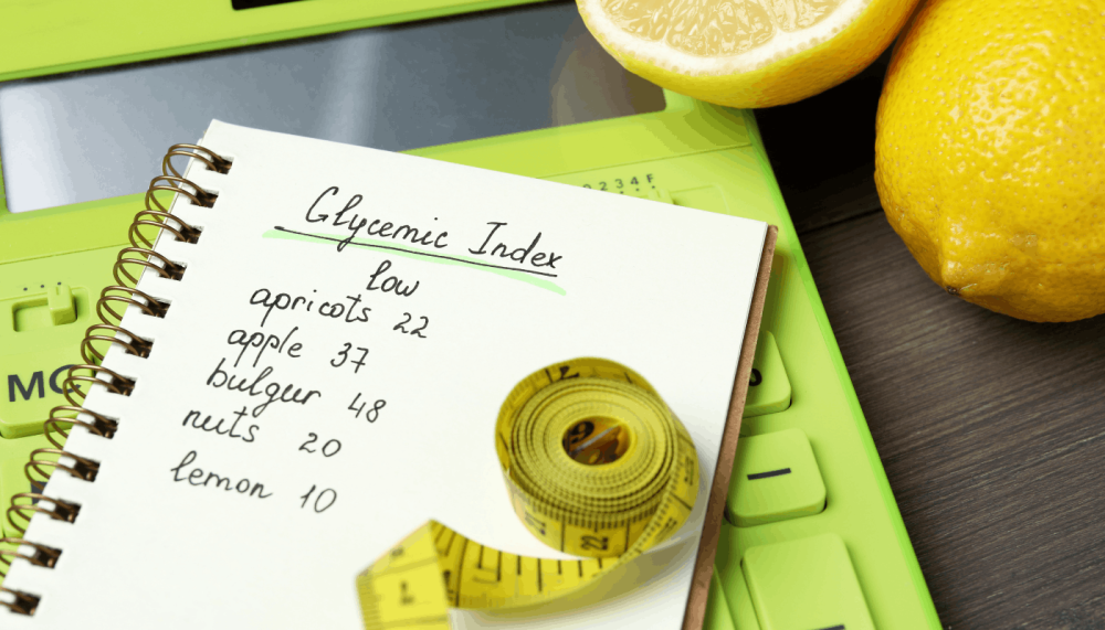 Dieta z niskim indeksem glikemicznym. Zbliżenie na otwarty notes z odręcznie wypisaną listą produktów i ich niskim indeksem glikemicznym (apricots 22, apple 37, bulgur 48, nuts 20, lemon 10), leżący obok zielonego kalkulatora i żółtej taśmy krawieckiej.